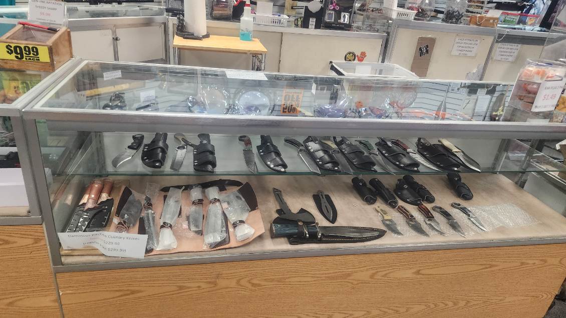 Glass display case of fixed-blade knives in leather belt sheaths above a row of Damascus culinary knives