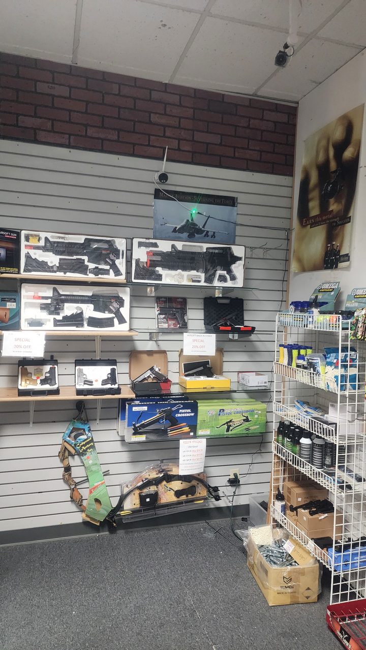 Store corner with airsoft rifles on the wall, pistol crossbows, a compound bow, and shelves of BBs and gas